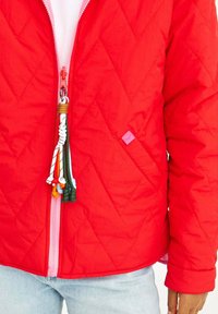 Červená prešívaná bunda s ružovým zipsom a farebnými strapcami. Pozoruhodný diamantový vzor a elastické manžety s jemným leskom. Denim viditeľný nižšie.