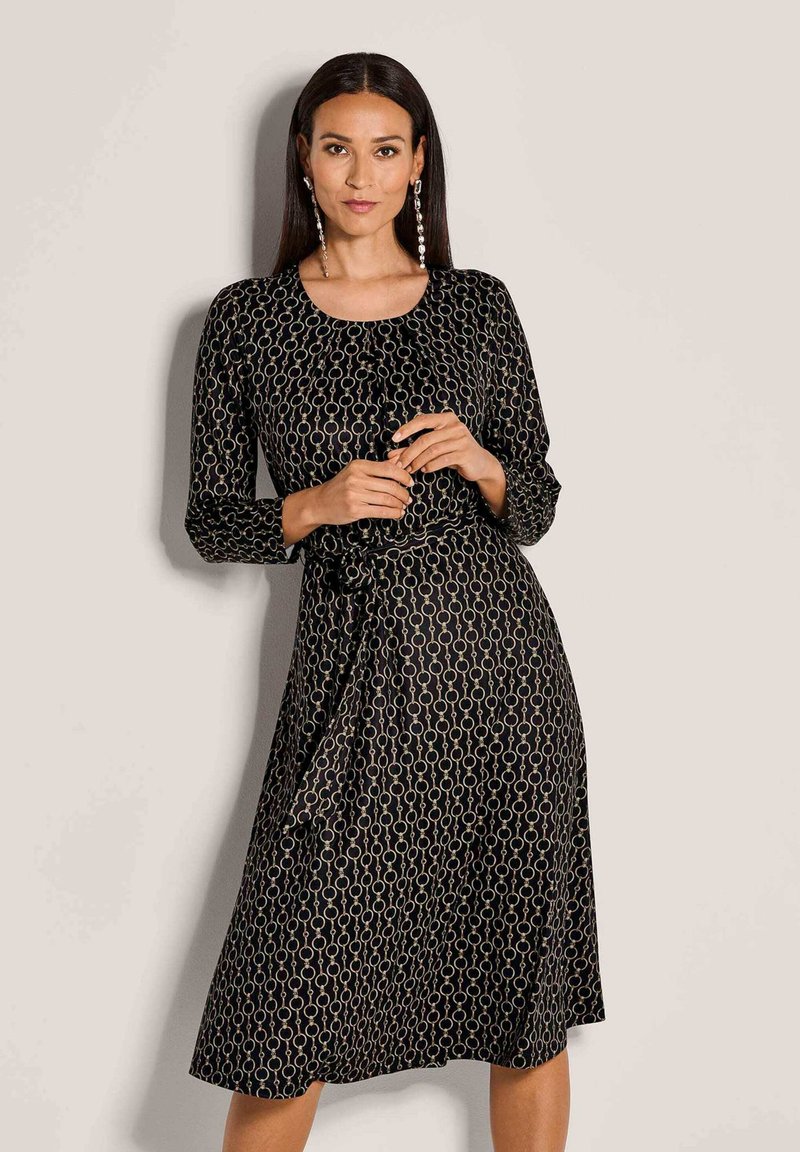 Frau mit langen dunklen Haaren, die ein schwarzes Midikleid mit kreisförmigem Kettenmuster und langen Ohrringen trägt, steht vor einem schlichten hellen Hintergrund.