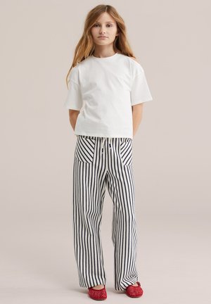 Jauna meitene stāv, valkājot vienkrāsainu baltu T-kreklu, melnas un baltas vertikālas svītras bikses ar priekšējām kabatām un sarkanas austas slīdēšanas kurpes.