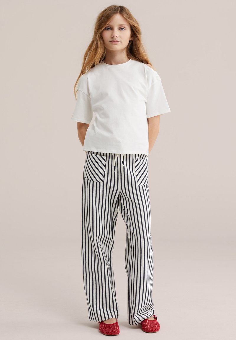 Jeune fille debout, portant un t-shirt blanc uni, un pantalon rayé vertical noir et blanc avec des poches avant, et des chaussures rouges tissées sans lacets.