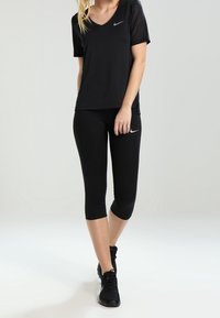 Mujer vistiendo una camiseta V de Nike negra, leggings recortados negros y zapatillas negras, de pie frente a un fondo claro y liso.