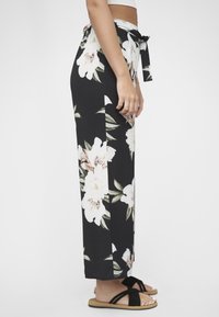 Svarte blomstrede bukser med store hvite blomster og grønne blader, med knyttet midje og avslappet passform, kombinert med svarte flate sandaler.
