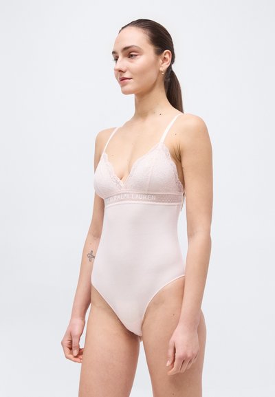 Νεαρή γυναίκα με αλογοουρά που φοράει κορμάκι ανοιχτό ροζ με δαντέλα, λεπτές τιράντες και λωρίδα με λογότυπο κάτω από το στήθος.