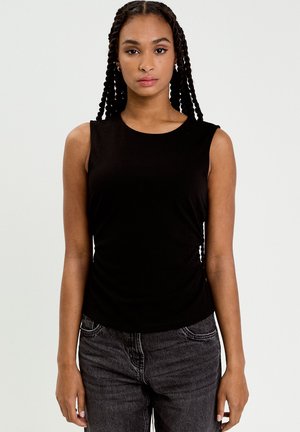 Femme aux cheveux tressés portant un haut noir sans manches et un jean gris foncé, debout devant un fond clair uni.