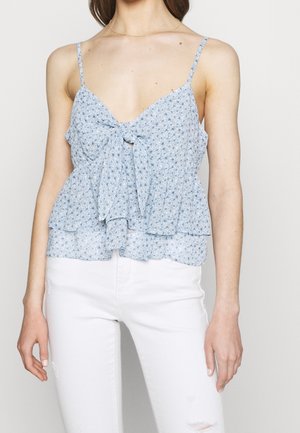 Vrouw draagt een lichtblauw bloemenmouwloos top met een knoop aan de voorkant en witte gescheurde spijkerbroek tegen een effen achtergrond.