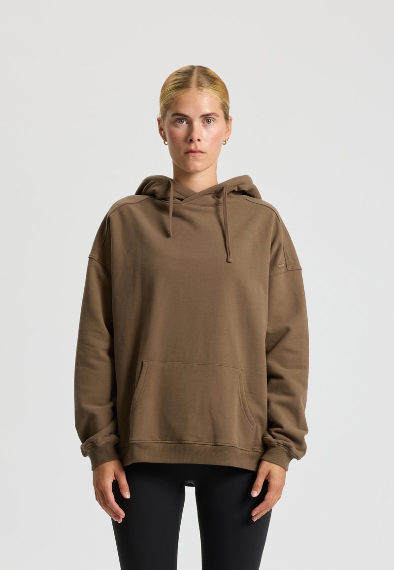 Felpa marrone realizzata in tessuto morbido, con una tasca frontale, cappuccio con cordino, vestibilità comoda e polsini a costine. Leggings neri sotto.