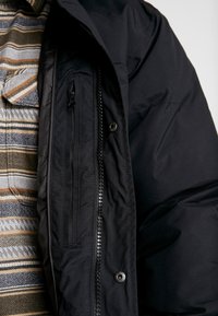 Black insulated jacket featuring a zippered pocket, textured fabric, and sleek design, layered over a multicolored plaid shirt.