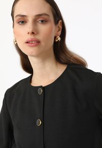 Veste noire à boutons, avec un col rond, ornée de quincaillerie dorée et d'une texture lisse. Le mannequin porte des boucles d'oreilles dorées et un maquillage naturel.