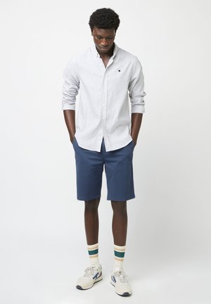 Homme debout les mains dans les poches, portant une chemise blanche à rayures longues, un short bleu marine, des chaussettes rayées et des baskets blanches sur fond blanc.