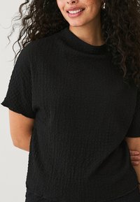 Zwarte top met structuur, korte mouwen en een hoge halslijn. Materiaal lijkt kreukelig met een zachte, licht verhoogde oppervlakte.