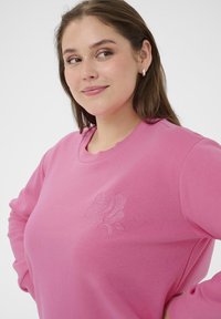 Sweatshirt rose en tissu doux avec une broderie de rose sur le côté gauche. Design col rond avec des manches longues et des poignets côtelés.