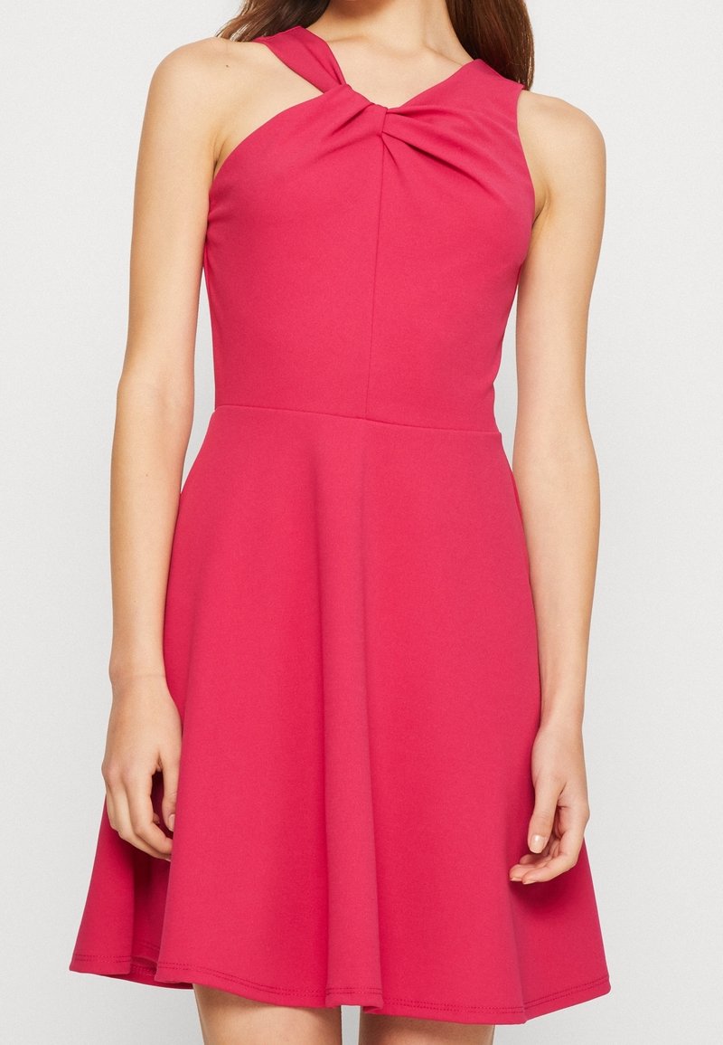 Robe rose sans manches avec un détail de nœud torsadé au niveau de l'encolure et une jupe évasée, portée par une femme debout, les bras détendus.