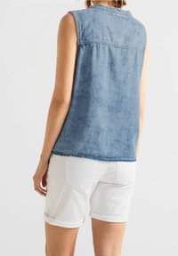 Sininen denimpaita ilman hihoja, jossa on hienovarainen haalistunut rakenne, suora leikkaus ja pyöristetty helma. Yhdistettynä valkoisiin käännettyihin shortseihin.