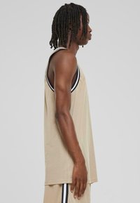 Beige ermeløs tanktop med mesh-tekstur, med sort kontrastkant langs halslinjen og armhulene, hengende løst.
