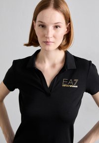 Camisa polo preta de mangas curtas, com um logotipo dourado "EA7 EMPORIO ARMANI" no lado esquerdo do peito e um design de gola clássica.