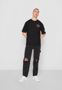 Black cotton t-shirt with front graphic in orange, paired with black ripped jeans and white sneakers. Simple, relaxed fit design.