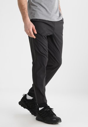 Hombre con pantalones deportivos negros, zapatillas negras, calcetines blancos y una camiseta gris, de pie frente a un fondo liso.