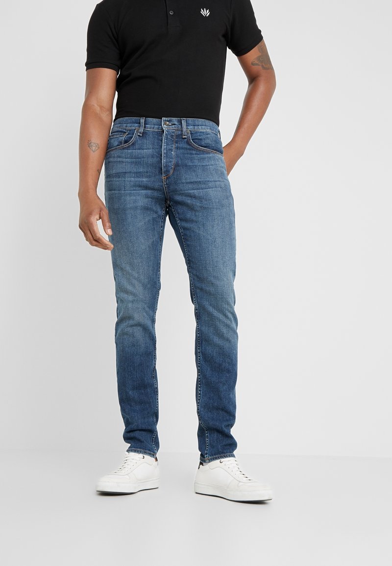 Pantalones vaqueros de hombre en azul, corte ajustado, diseño de tiro medio, ligero desvanecimiento, cinco bolsillos, combinados con zapatillas blancas y una camiseta polo negra.