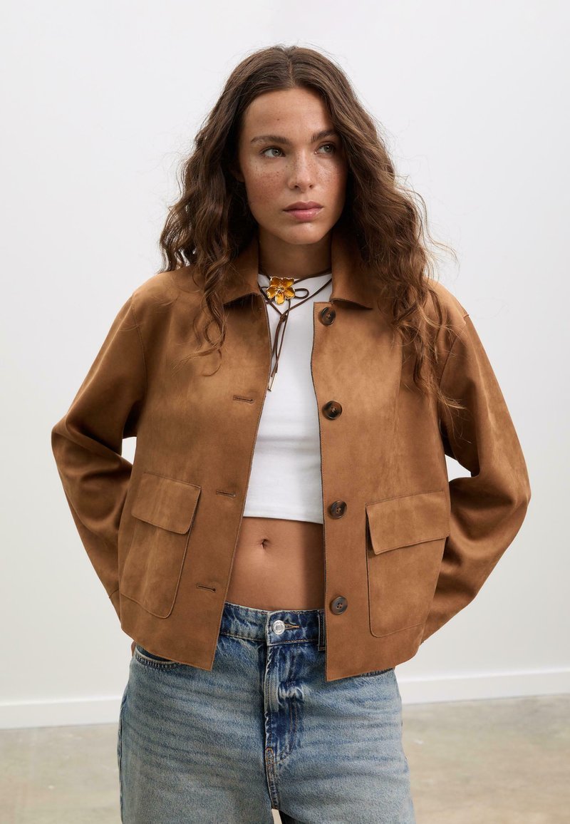 Veste courte en suède marron avec un col, fermeture à boutons et poches avant. Associée à un haut court blanc et un jean bleu déchiré.