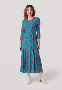 Turquoise lange mouw jurk met een bloemenpatroon in roze en rood, met een tailleband en een gelaagde rok; gecombineerd met witte haklaarzen.