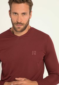 Camicia a maniche lunghe color bordeaux con scollo a V, realizzata in tessuto liscio. Presenta un logo discreto nell'angolo superiore sinistro.
