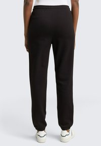 Schwarze Sweatpants mit elastischem Bund und bündchenförmigen Knöcheln, die eine glatte Textur und einen lockeren Schnitt aufweisen. Getragen mit weißen Turnschuhen.