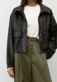 Giacca cropped in pelle nera con chiusura con zip, tasche laterali e polsini elasticizzati, abbinata a pantaloni olive verdi a vita alta.