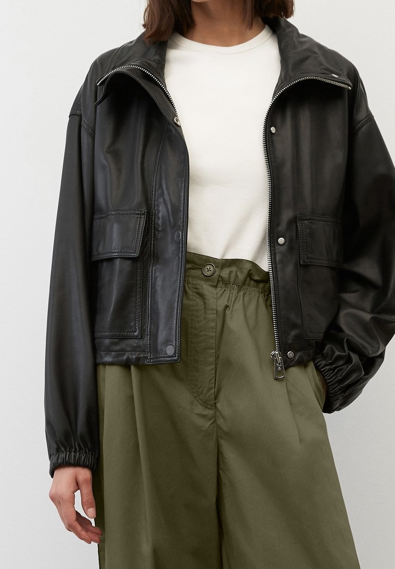 Giacca cropped in pelle nera con chiusura con zip, tasche laterali e polsini elasticizzati, abbinata a pantaloni olive verdi a vita alta.