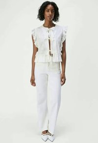 White cotton top with eyelet detailing, ruffled sleeves, and a tie front. Paired with white pants and pointed white shoes.