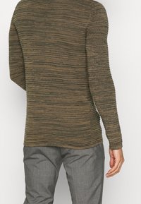 Langärmliger Pullover in einem strukturierten braun-und-grünen Streifenmuster. Figurbetontes Design mit gerippten Kanten. Kombiniert mit grauen Hosen.