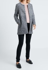 Szary, teksturowany płaszcz z okrągłym kołnierzem, otwarty z przodu, z dwoma bocznymi kieszeniami, noszony na jasnoróżowym swetrze i czarnych dopasowanych spodniach.