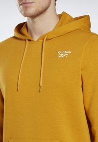 Sweat à capuche jaune moutarde en tissu doux, avec une poche kangourou, une capuche à cordon de serrage, et un logo Reebok blanc sur la poitrine.