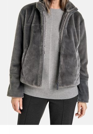 Personne portant une veste zippée en fausse fourrure grise par-dessus un pull en maille gris clair et un pantalon gris foncé, debout devant un fond uni.