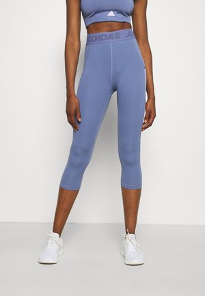 Kvinde iført cropped blå Adidas leggings og matchende sports-bh med hvide sneakers, stående mod en ensfarvet baggrund.