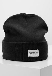 Crna pletena kapa s preklopom, koja ima bijelu pravokutnu oznaku s nazivom brenda "oxmo" u crnom tekstu. Mekana tekstura.