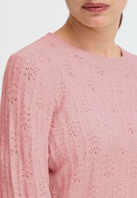 Maglione lavorato a maglia rosa chiaro con una morbida trama, caratterizzato da un motivo decorativo di punti traforati e un classico scollo rotondo.