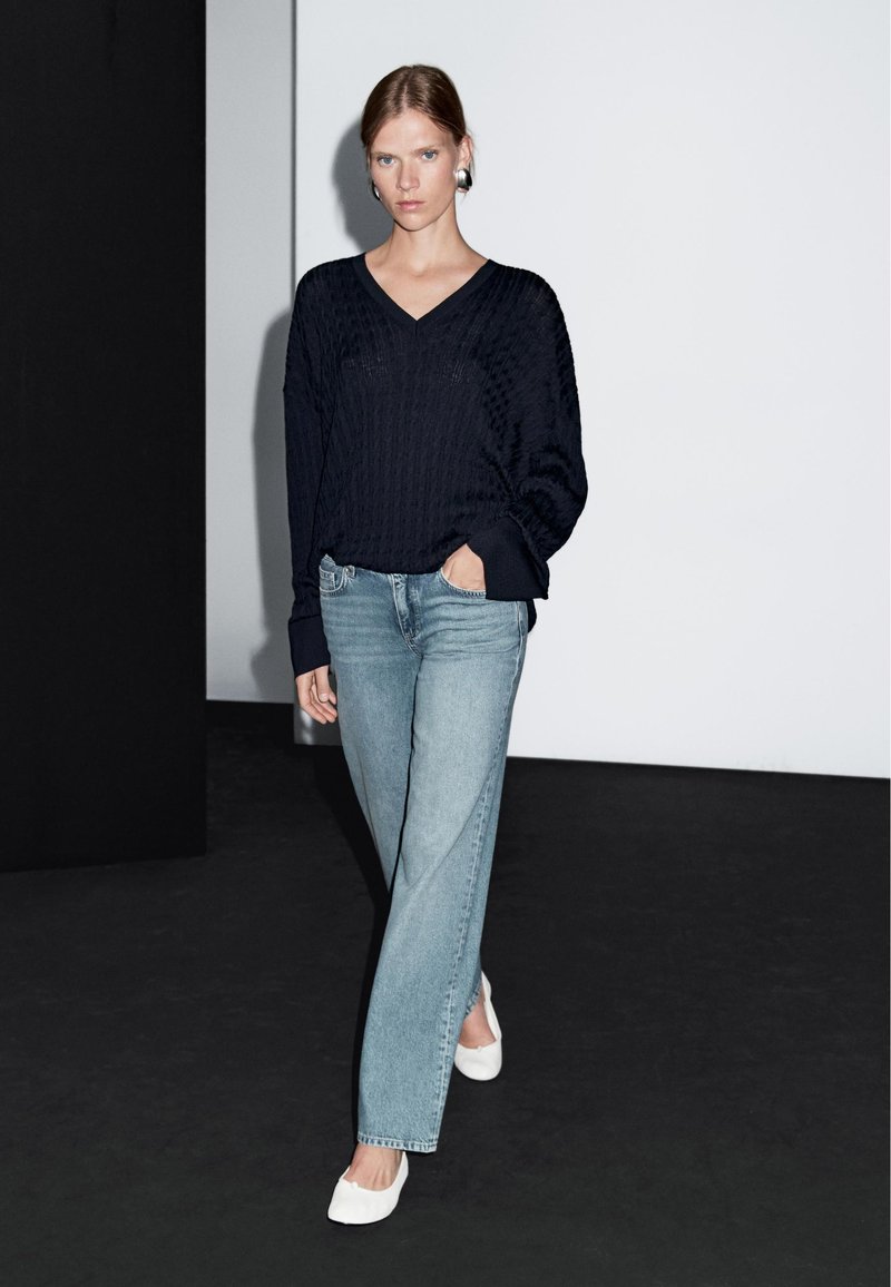 Marinblå stickad tröja med V-ringning, ihop med ljusblå raka jeans och vita lägenheter. Outfiten har en avslappnad passform och ribbad textur.