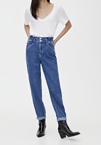 Vaqueros de mezclilla azul de cintura alta con cintura fruncida, dos bolsillos frontales y dobladillos con vuelta, combinados con una camiseta blanca de cuello en V y botas negras.