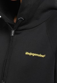Czarna bluza z kapturem wykonana z miękkiego materiału, z żółtym logo "khujo genuine" i regulowanym sznurkiem w kołnierzu z oznakowaniem.