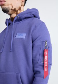 Fialová flísová mikina s klokankou, kapucňou so šnúrkou a zipsovou vreckom na rukáve so červeným označením „REMOVE BEFORE FLIGHT“. 