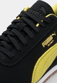 Gros plan sur une paire de sneakers Puma Roma noir et jaune, montrant les lacets noirs, une bande jaune cousue, et le logo doré Puma Roma sur le côté.