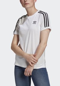 Balta medvilninė T-shirt maikė su juodu Adidas logotipu, juoda trijų juostelių apdaila ant rankovių, suktos rankovės ir laisvas siluetas. Su derinami mėlyni džinsai.