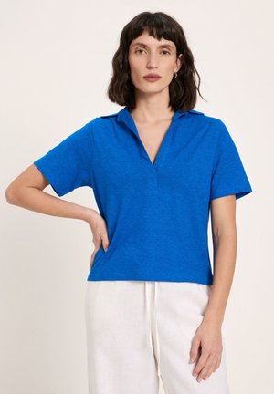 Frau mit dunklen Haaren trägt ein leuchtend blaues Poloshirt mit kurzen Ärmeln und weiße Hosen mit Kordelzug, steht mit einer Hand in der Hüfte.