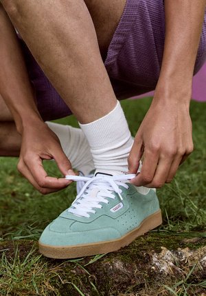 Handen die witte veters strikken aan een lichtgroene sneaker met een zool van schoenzoolrubber, persoon draagt witte sokken en paarse korte broek, buiten op gras en schors.