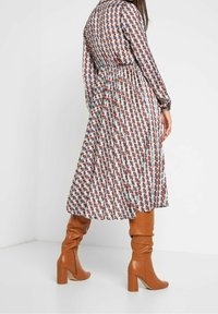 Šaty s dlouhým rukávem v geometrickém vzoru červené, modré a bílé barvy, se staženým pasem. V kombinaci s hnědými kozačkami na vysokém podpatku po kolena.