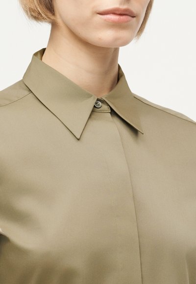 Κοντινό πλάνο ενός ατόμου που φοράει μπεζ πουκάμισο με κουμπιά και μυτερό γιακά, με κοντά ανοιχτό καφέ μαλλιά.