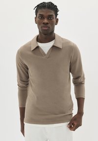 Joven con el cabello corto y rizado, vistiendo una camisa de manga larga beige con cuello sobre una camiseta blanca y pantalones blancos, contra un fondo neutro.