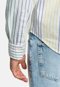 Gestreifter Baumwollhemd mit grünen, blauen und gelben Streifen, mit Knopfmanschetten. Kombiniert mit hellblauen Jeans.