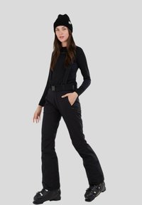 Schwarzes Skianzug mit verstellbaren Schultergurten, enganliegendem Langarmshirt und Schneehose. Kombiniert mit schwarzen Skischuhen und einer Mütze.