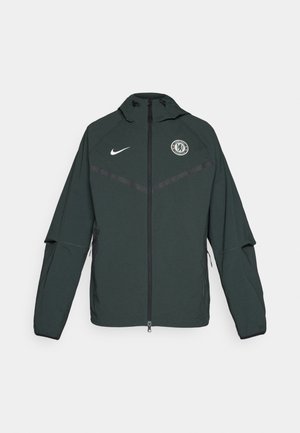 Giacca Nike verde scuro con cappuccio, zip frontale, logo del Chelsea FC sul petto sinistro e strisce orizzontali nere a contrasto.